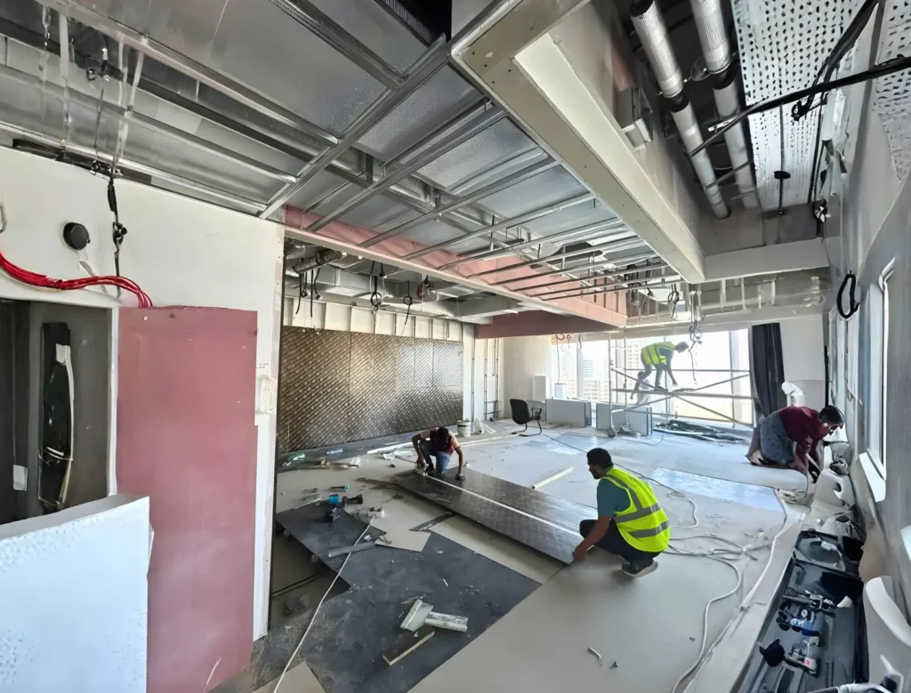 Several construction workers in safety vests are installing panels and working with tools inside a partially finished building with exposed pipes, wires, and ceiling framework. Debris and construction materials are scattered on the floor.