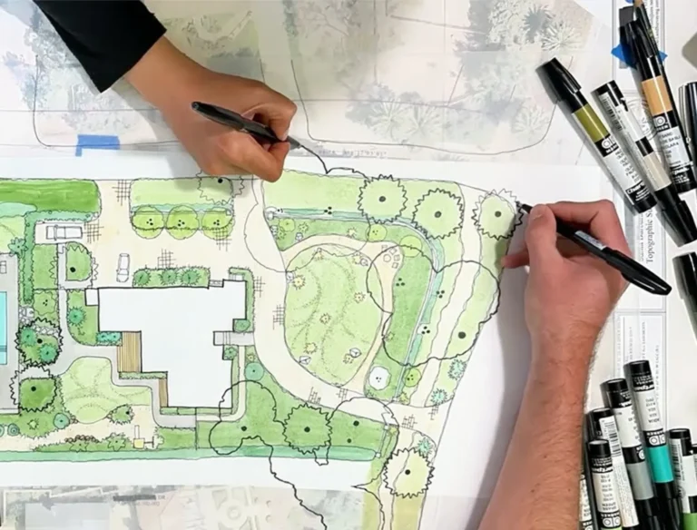 Two people are drawing on a landscape architecture plan using black pens. The plan features green areas, trees, and pathways. Several colored markers are scattered on the table around the drawing.
